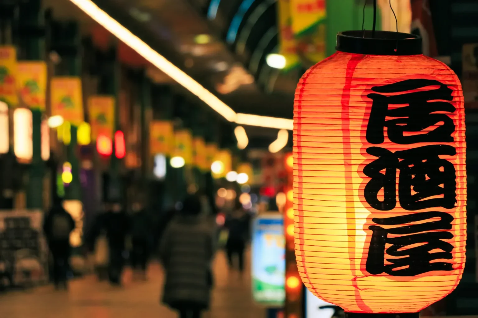 A glowing red paper lantern with the word izakaya brushed on it in black, hanging in a shopping arcade with blurred shop signs and people behind