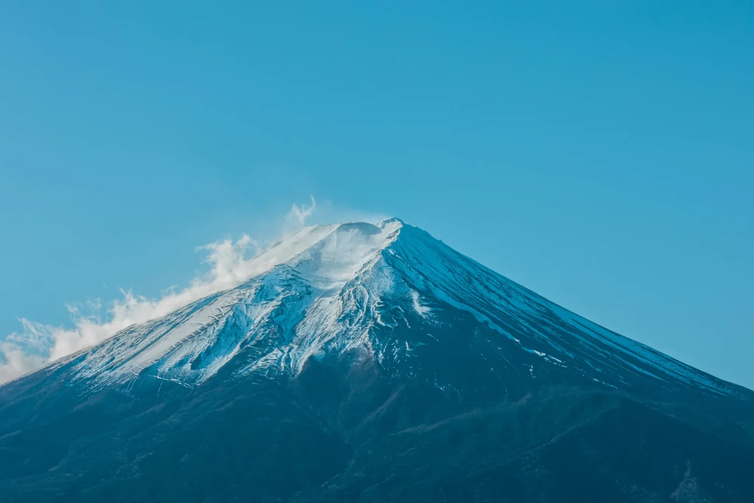 Snow-covered summit of Mount Fuji against a clear blue sky, with wind lifting snow off the top, seen from the Fujinomiya side in Shizuoka