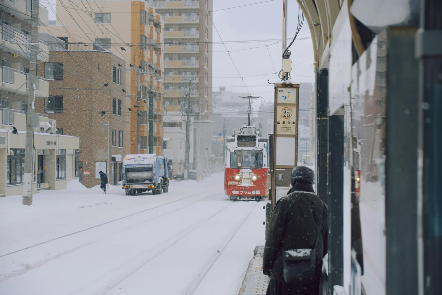 A red Sapporo streetcar approaching the Yamahana 9-jo stop on a snow-covered street, with a passenger waiting under the shelter