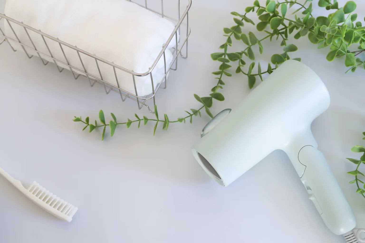 A pale green hair dryer lying on a white surface beside a comb, a wire basket with a rolled white towel and green sprigs