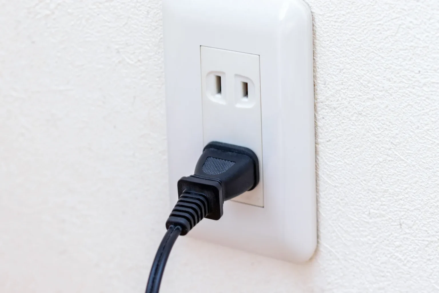 A black plug pushed into the lower socket of a white two-socket wall outlet, its cable hanging down