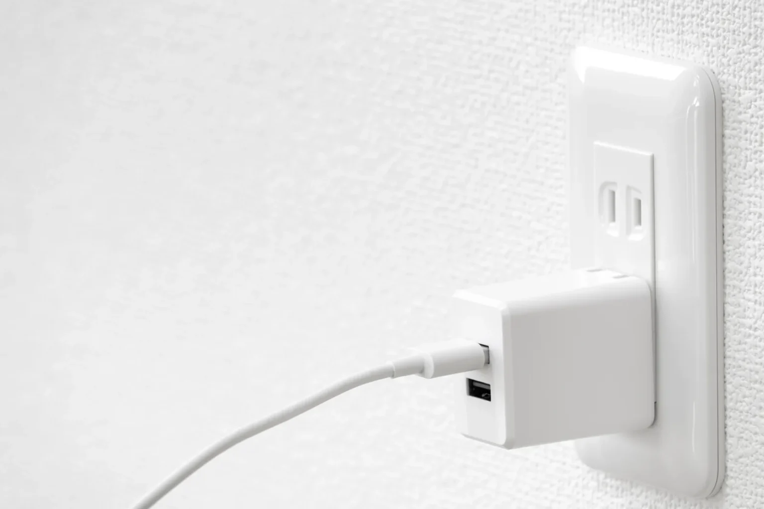 A white USB charger with a cable attached, plugged into a white wall outlet on a textured white wall