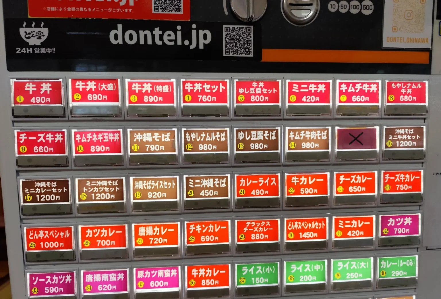 Close-up of a restaurant meal-ticket machine with rows of red, brown, orange, pink and green buttons, each showing a dish name in Japanese and a price in yen, such as gyudon 490 yen, with coin slots marked 10, 50, 100 and 500 at the top and one button covered with a cross