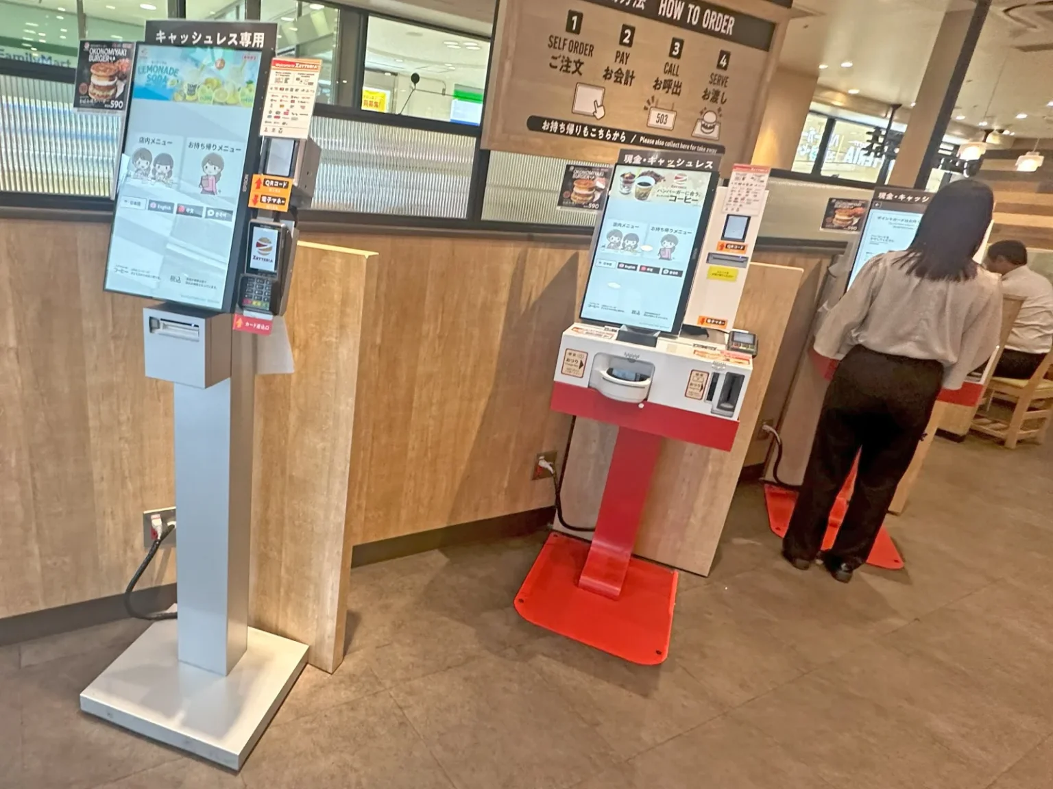 Self-order kiosks at a fast-food restaurant: on the left a touchscreen on a grey stand under a sign reading cashless only in Japanese, in the centre a red kiosk with a coin tray under a sign reading cash and cashless, and a wooden board above showing four steps: self order, pay, call, serve