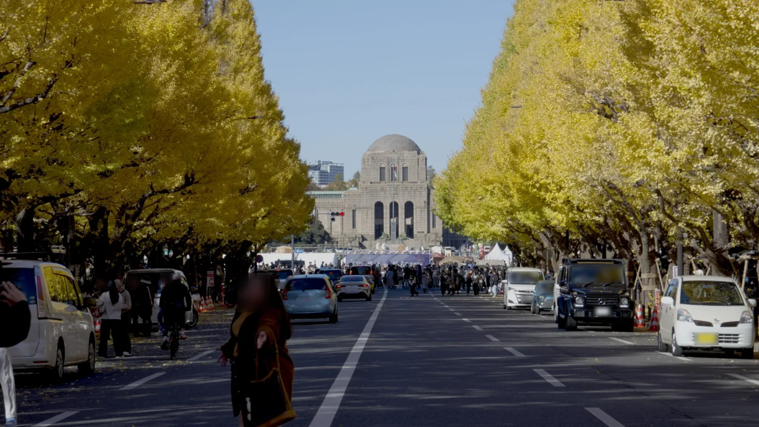 The Meiji Jingu Gaien ginkgo avenue in Tokyo looking toward the gallery, with cars parked along both kerbs, traffic cones, people and a cyclist on the road, and white marquees and a mass of people under a banner in front of the domed Meiji Memorial Picture Gallery
