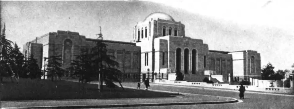 Black-and-white photograph of the Meiji Memorial Picture Gallery in Tokyo, a stone building with a central dome and tall arched openings, seen across a lawn with conifers and a few people walking on a curved drive