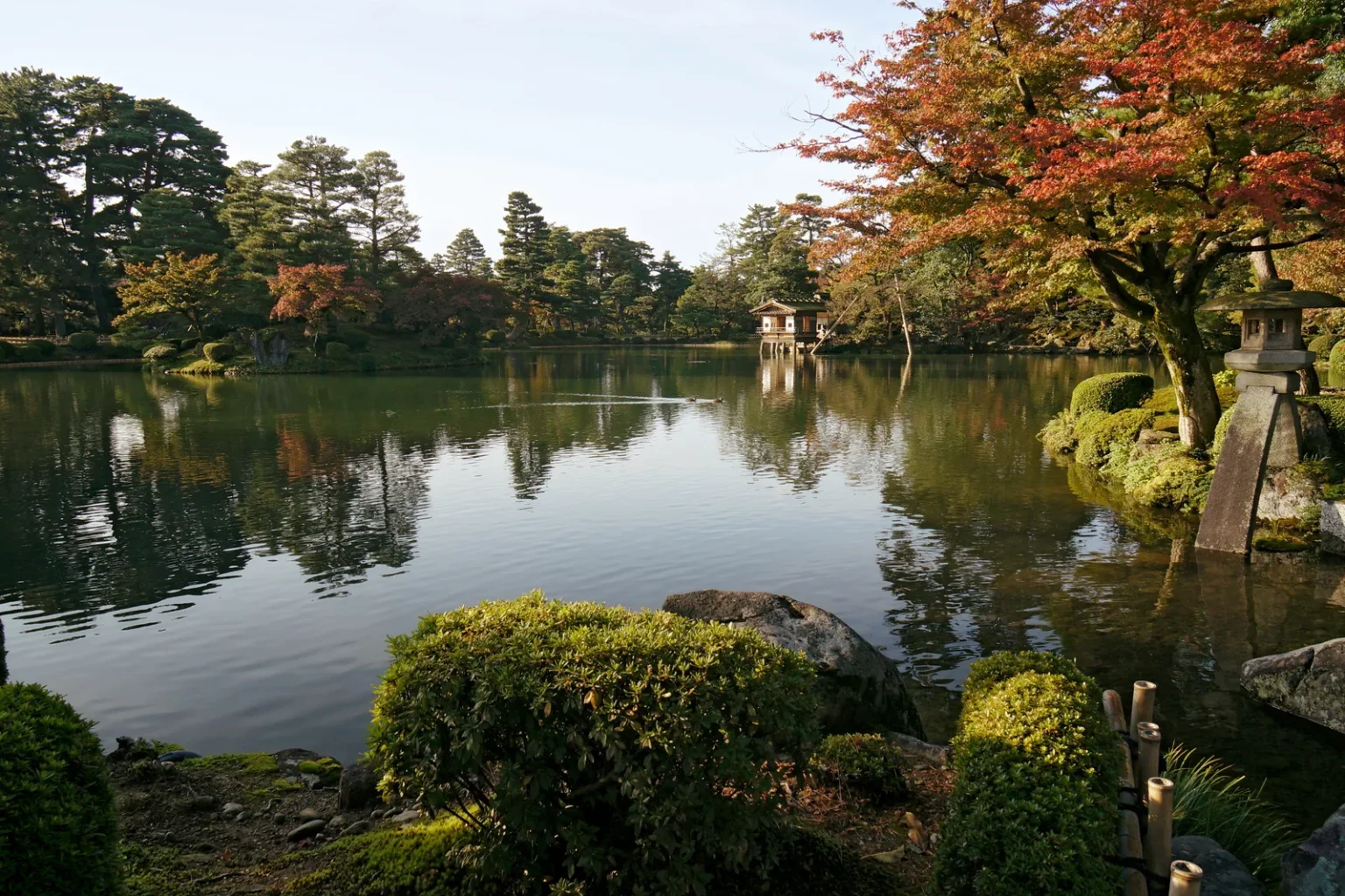 Kasumigaike pond at Kenrokuen reflecting tall pines and trees, with a maple turning red and orange on the right above a two-legged stone lantern at the water’s edge, a small pavilion on the far shore, and clipped shrubs in the foreground