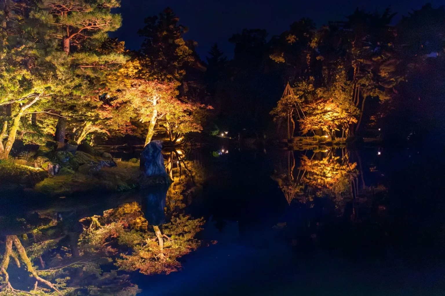 Kenrokuen at night with lit trees in yellow, orange and green reflected in a dark pond, a large rock at the water’s edge and, on the right, a cone of poles and ropes lit above the shore