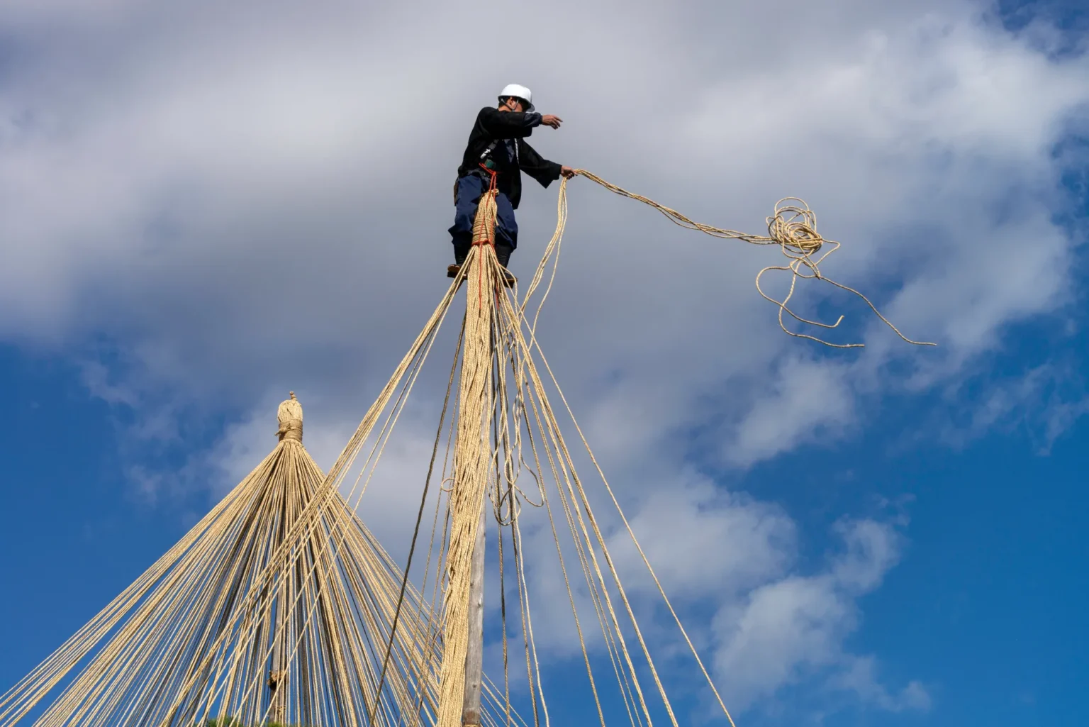 A worker in a white helmet stands at the top of a tall pole against a blue sky with clouds, throwing out a pale rope, while dozens of ropes run down from the pole and from a second finished cone of ropes on the left
