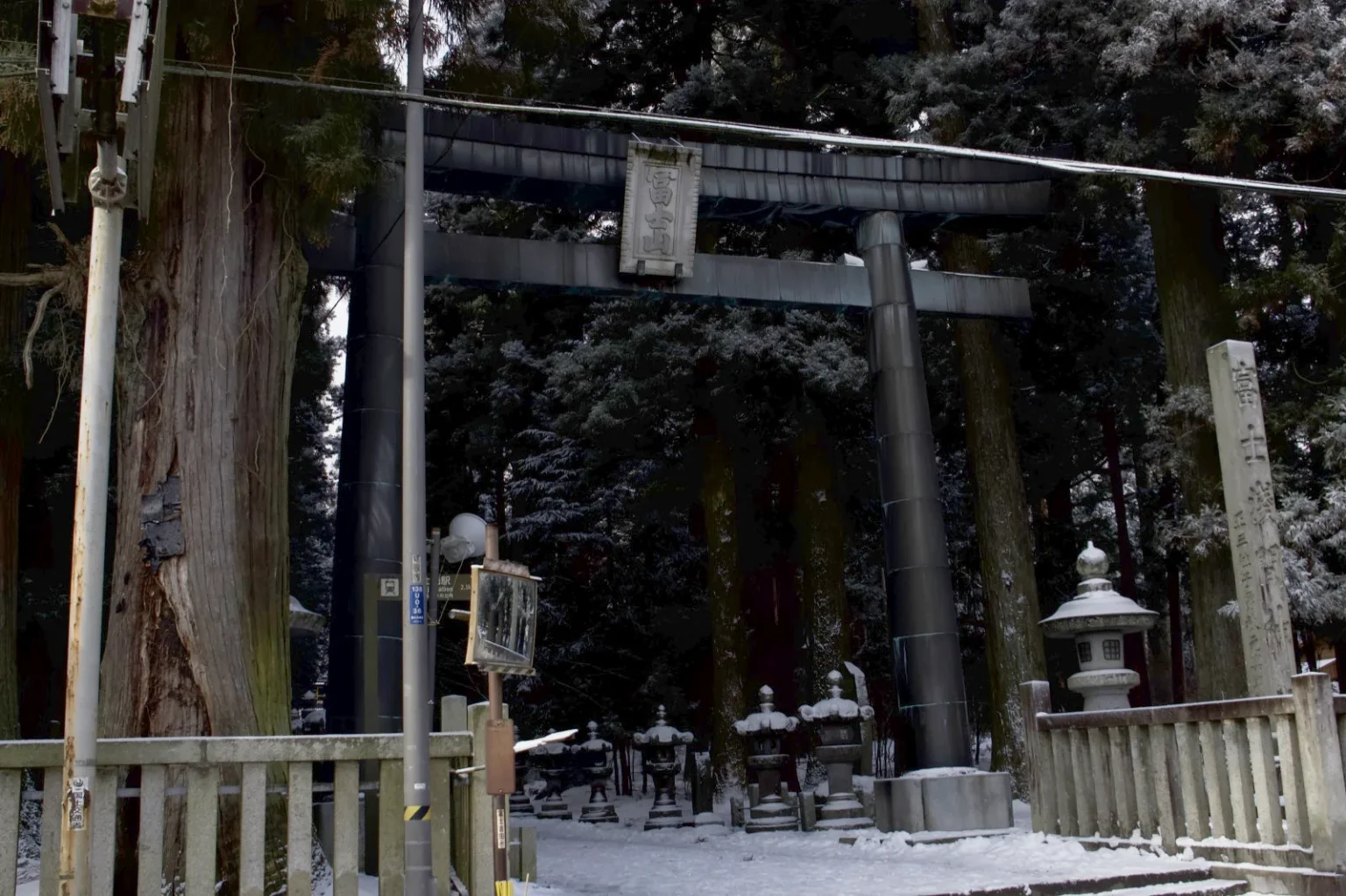 A dark-coloured torii with a stone plaque reading 冨士山 at the entrance to the shrine approach, low stone fences on both sides, snow-covered stone lanterns and cedars beyond it, a thick cedar trunk and a utility pole on the left and a tall inscribed stone post on the right, at 8:19 a.m. on 9 March 2025