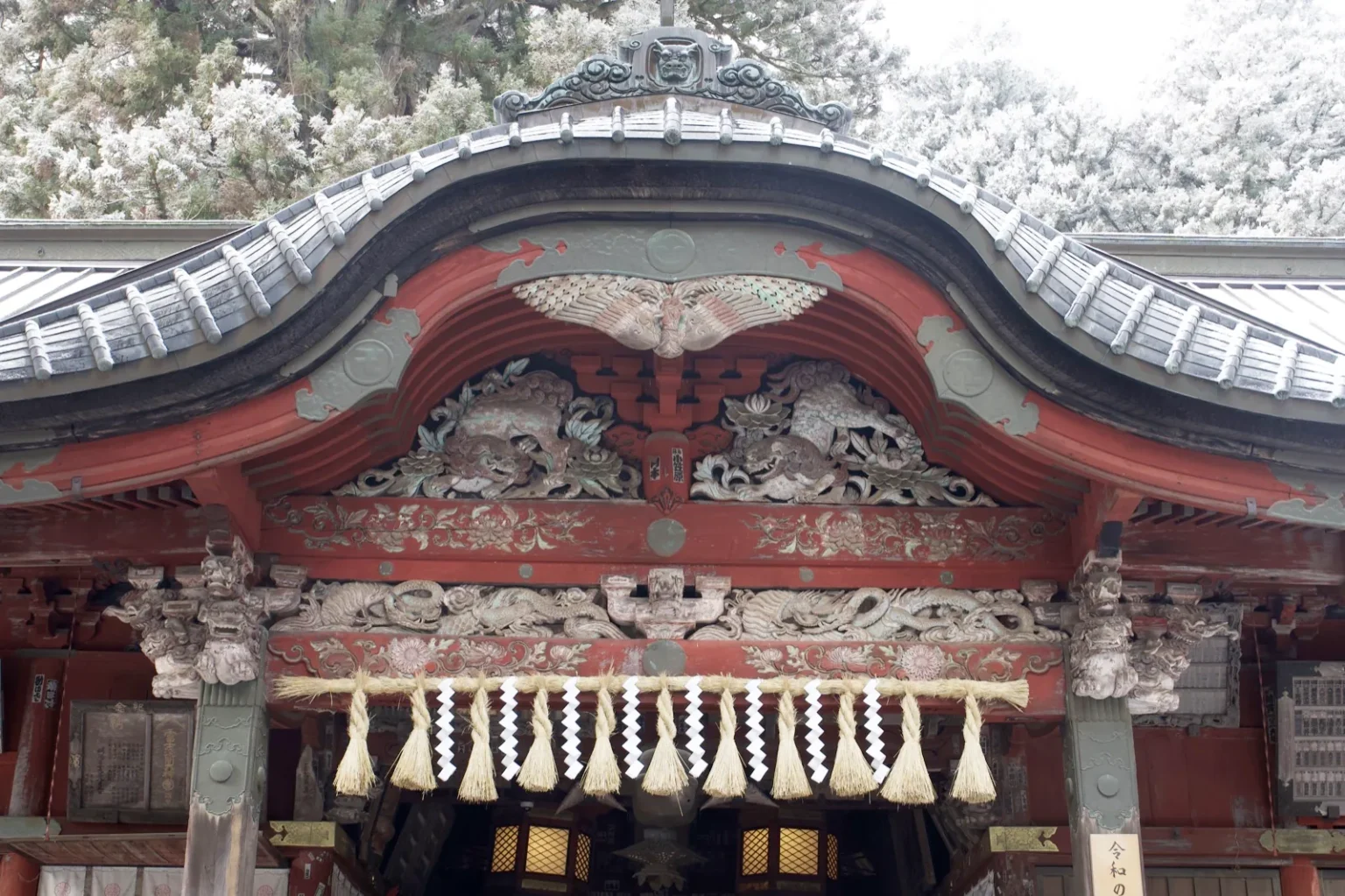 Close view of the curved kara-hafu gable over the entrance of the worship hall, painted red with carved and coloured panels below the roof, a thick straw rope with white paper streamers hanging across the front, and snow-covered trees behind the roof, at 7:54 a.m. on 9 March 2025
