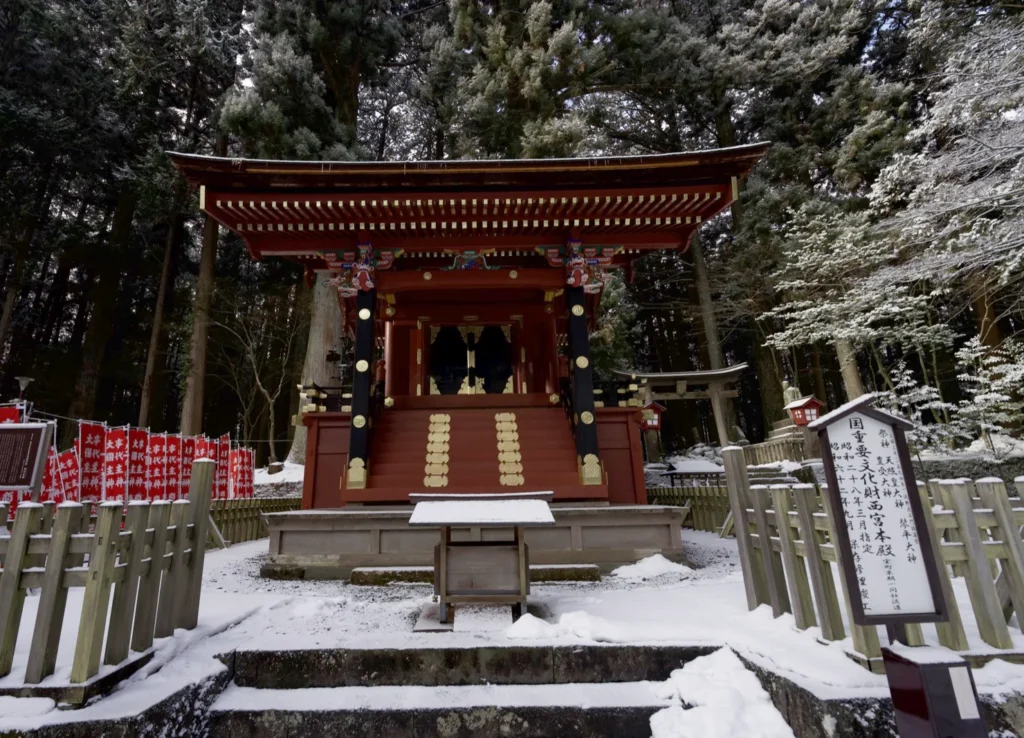 The small red Nishimiya Honden sanctuary with black pillars and gold fittings behind a wooden fence, a white signboard on the right reading 国重要文化財 西宮本殿, rows of red banners at left, and behind the fence to the right a plain wooden torii with red lantern boxes and stone steps, all under snow-covered cedars, at 8:02 a.m. on 9 March 2025