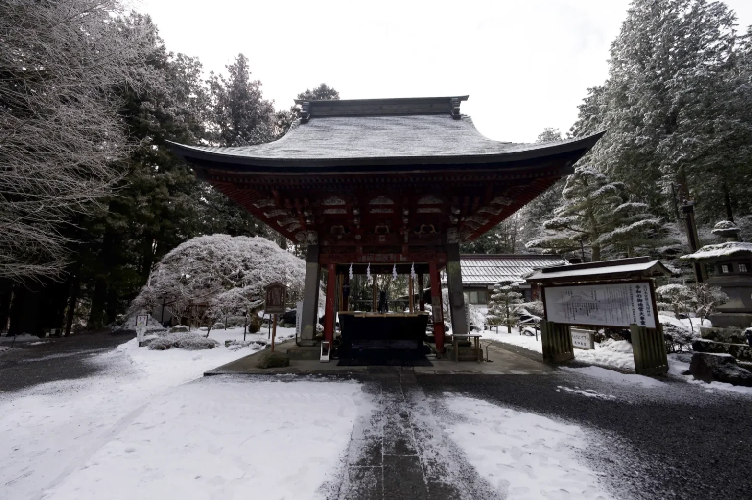The temizuya water pavilion of Kitaguchi Hongu Fuji Sengen Shrine, a red-pillared open structure with a large grey roof with upswept eaves, a stone basin underneath, a signboard to its right, snow on the ground and snow-covered trees behind, at 7:58 a.m. on 9 March 2025