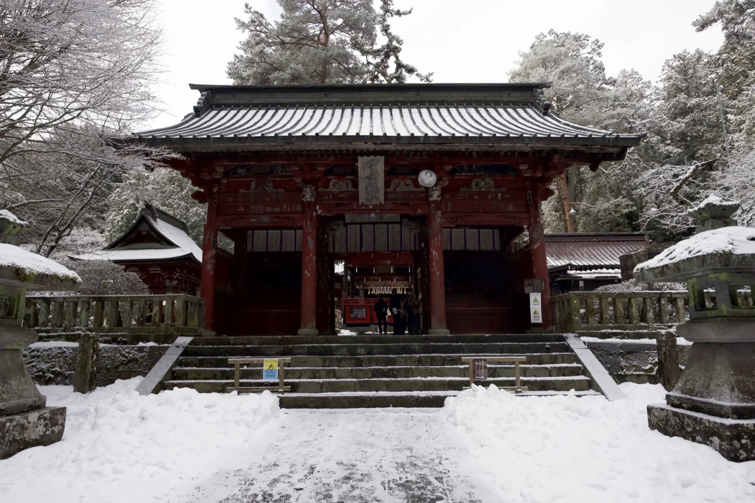 The red Zuishinmon gate of Kitaguchi Hongu Fuji Sengen Shrine with a grey ribbed copper roof dusted with snow, stone steps leading up to it, stone lanterns at both sides and snow piled along the path, at 8:10 a.m. on 9 March 2025
