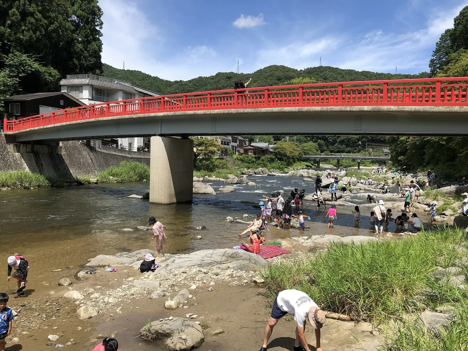 Summer scene: people wading and sitting on rocks in a shallow river under a red-railed concrete bridge, with green trees and hills behind and a second bridge in the distance, at Korankei