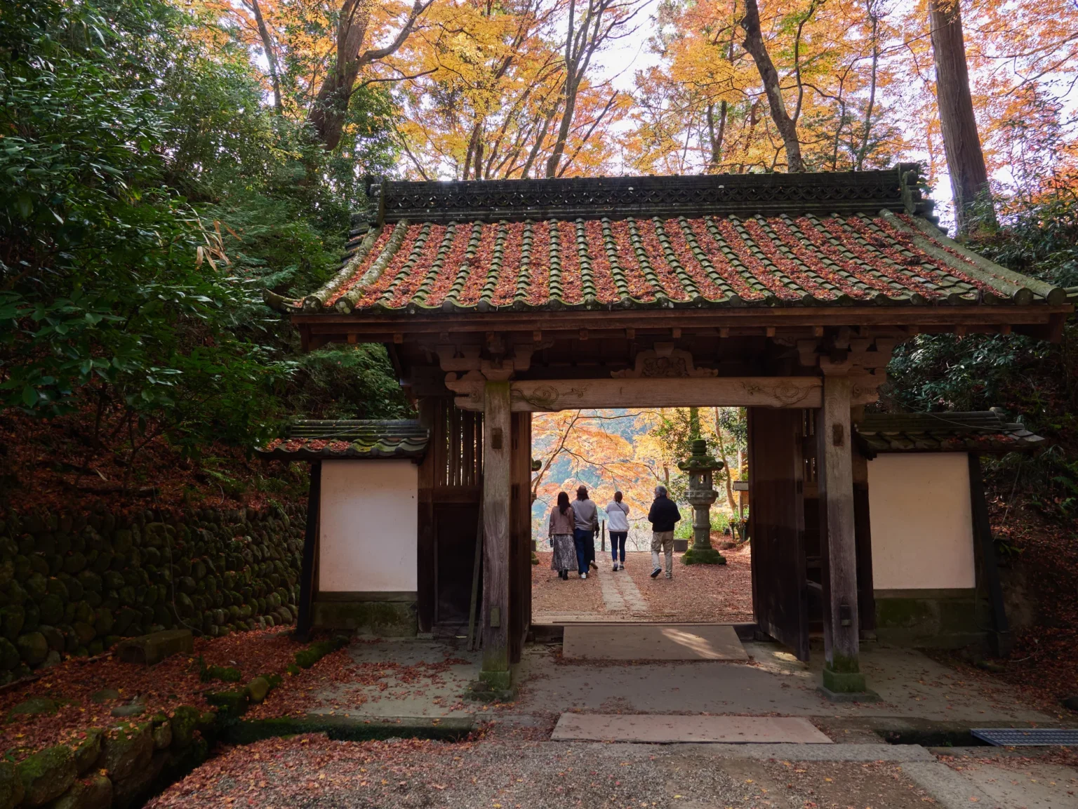 A wooden temple gate with a tiled roof and white plaster side walls, framing four people walking along a leaf-covered stone path towards a stone lantern under orange and yellow trees