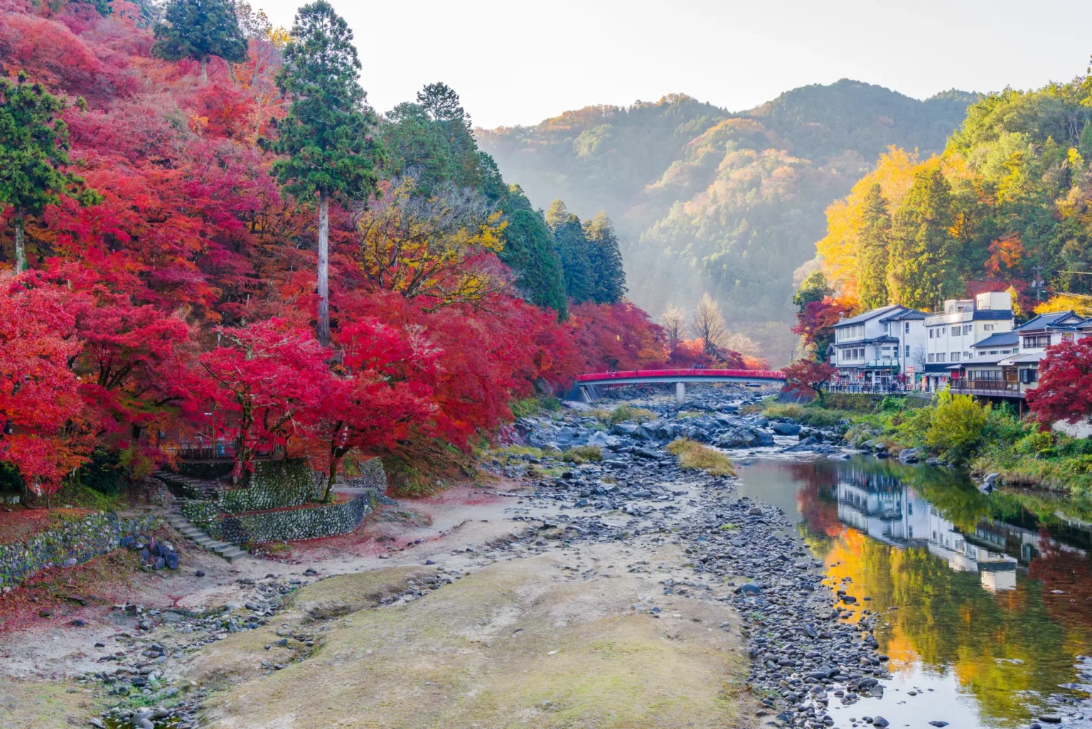 Red and orange maples covering the left bank of a shallow, stony river, with a red-railed bridge crossing it in the middle distance, white buildings and yellow trees on the right bank reflected in the water, and misty forested hills behind