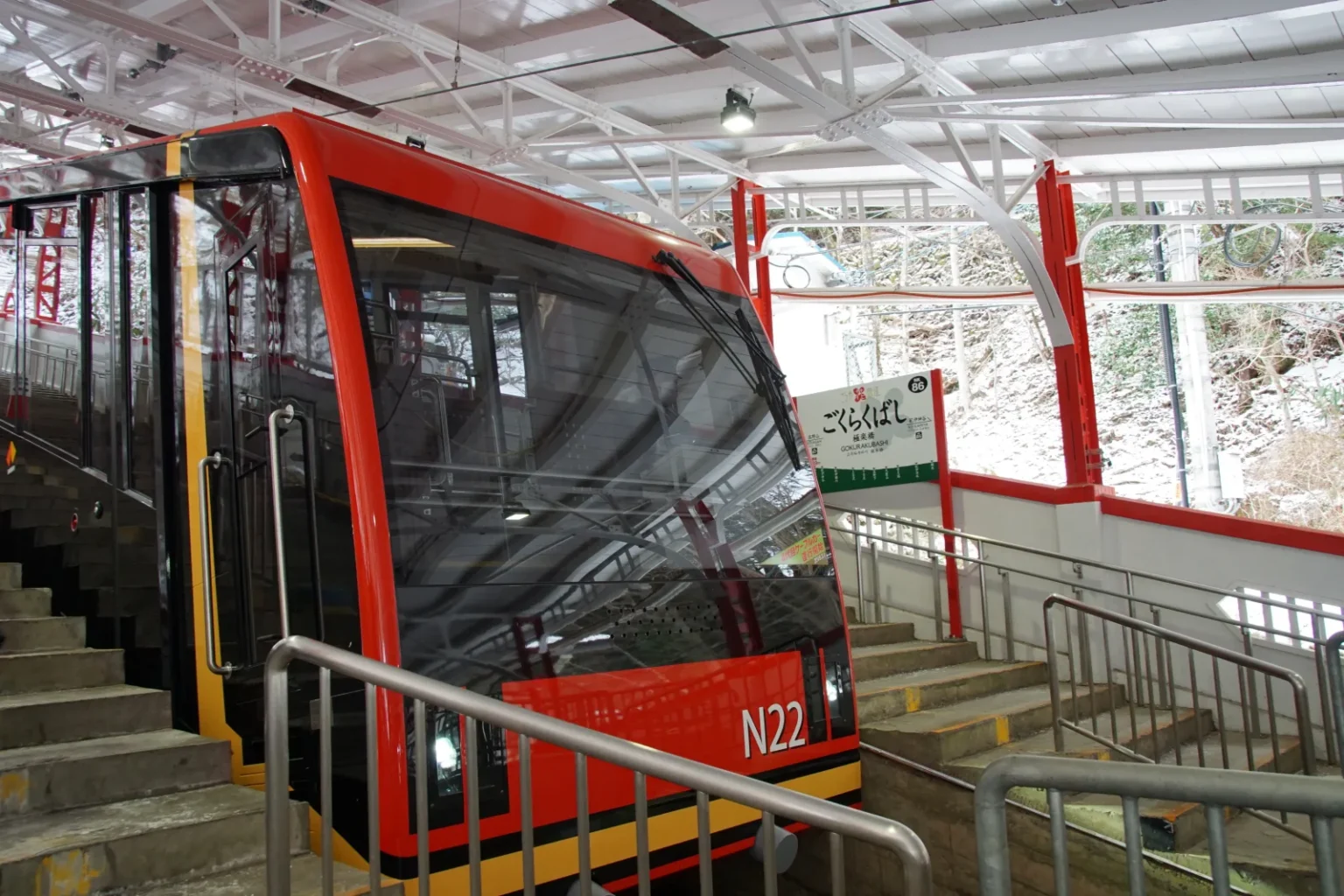 A red Koyasan cable car standing at the stepped platform of Gokurakubashi station under a white steel roof, with the station name sign behind and snow on the slope