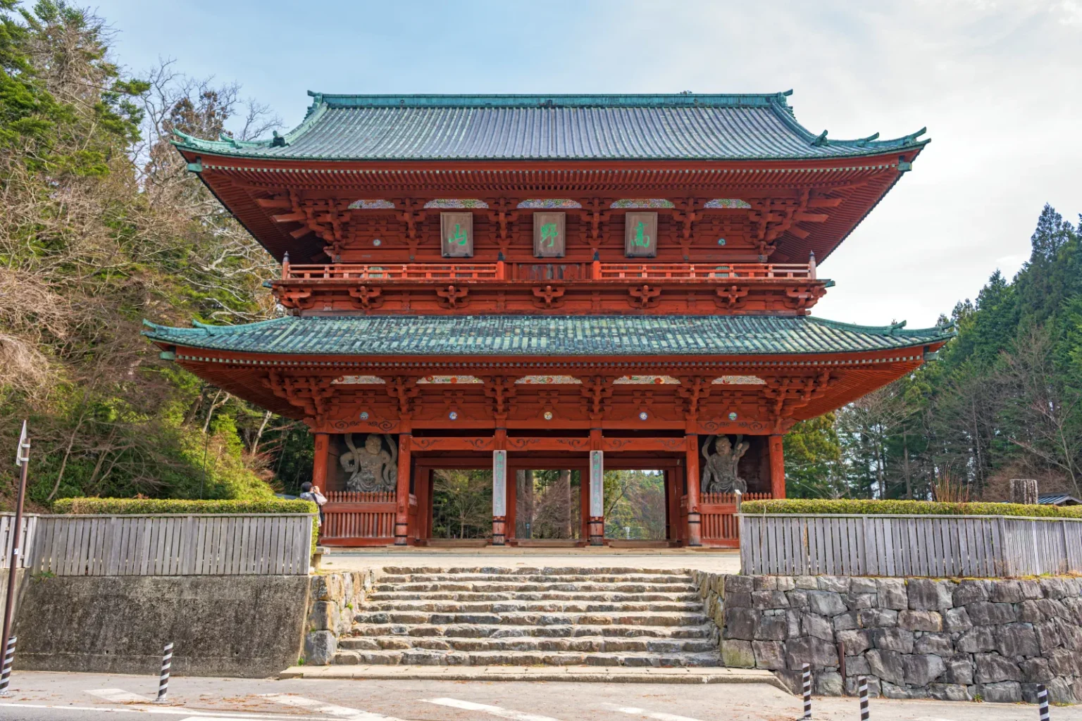 The tall red two-storey Daimon gate at the western entrance to Mount Koya, at the top of stone steps with forest on the hillside