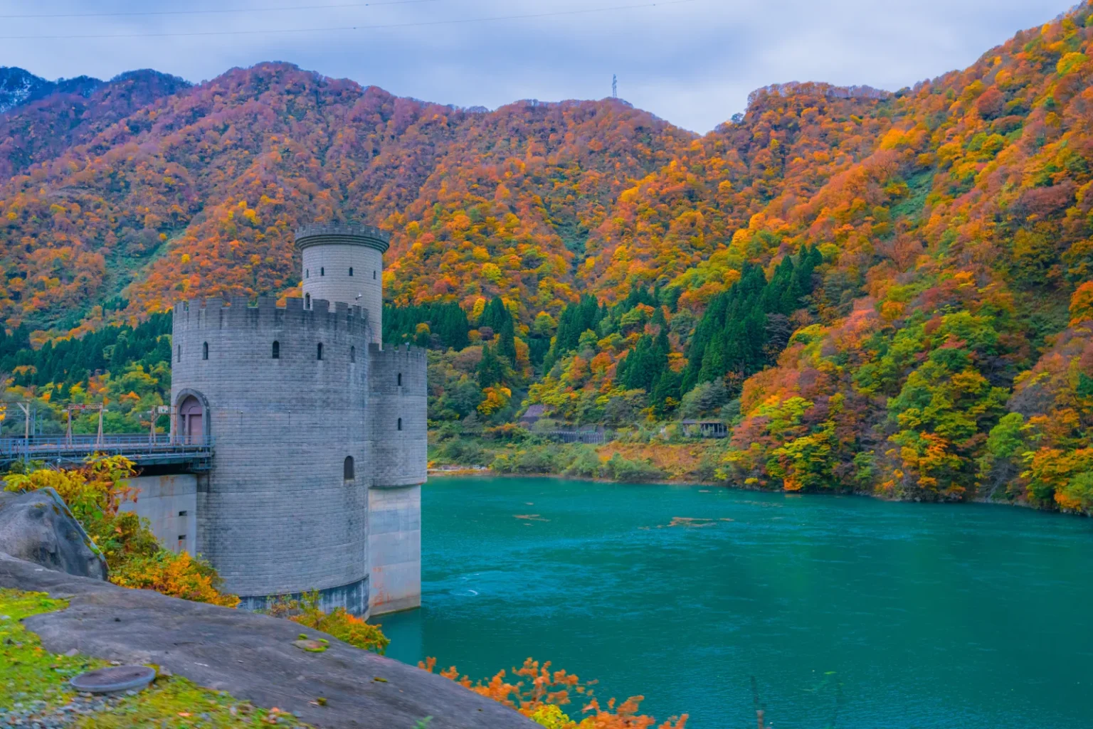 A grey stone tower building at the edge of a turquoise reservoir, below mountainsides covered in orange, red and yellow autumn trees