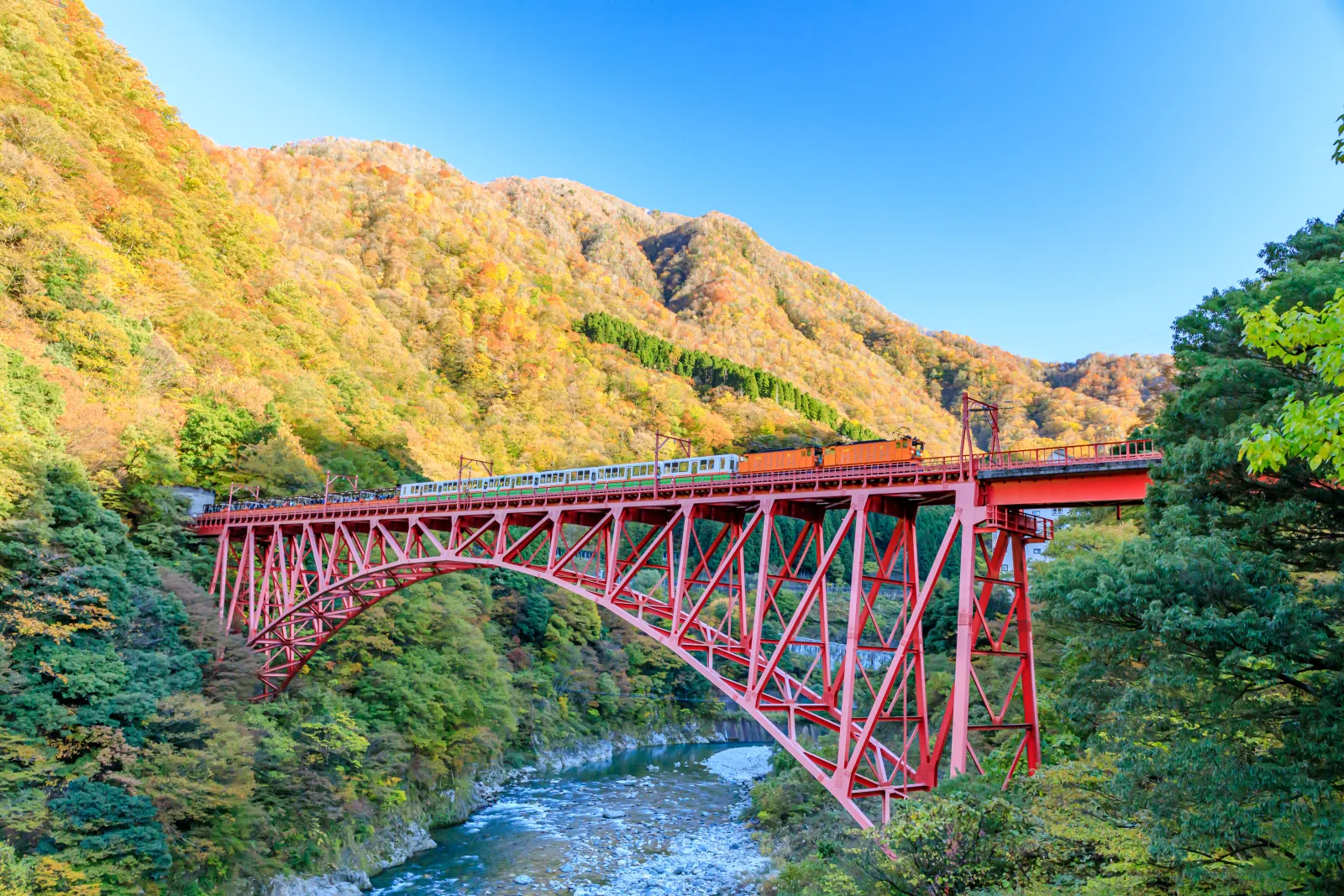 A Kurobe Gorge Railway train with orange locomotives crossing a red steel arch bridge above a blue-green river, with mountainsides in yellow and orange autumn colour