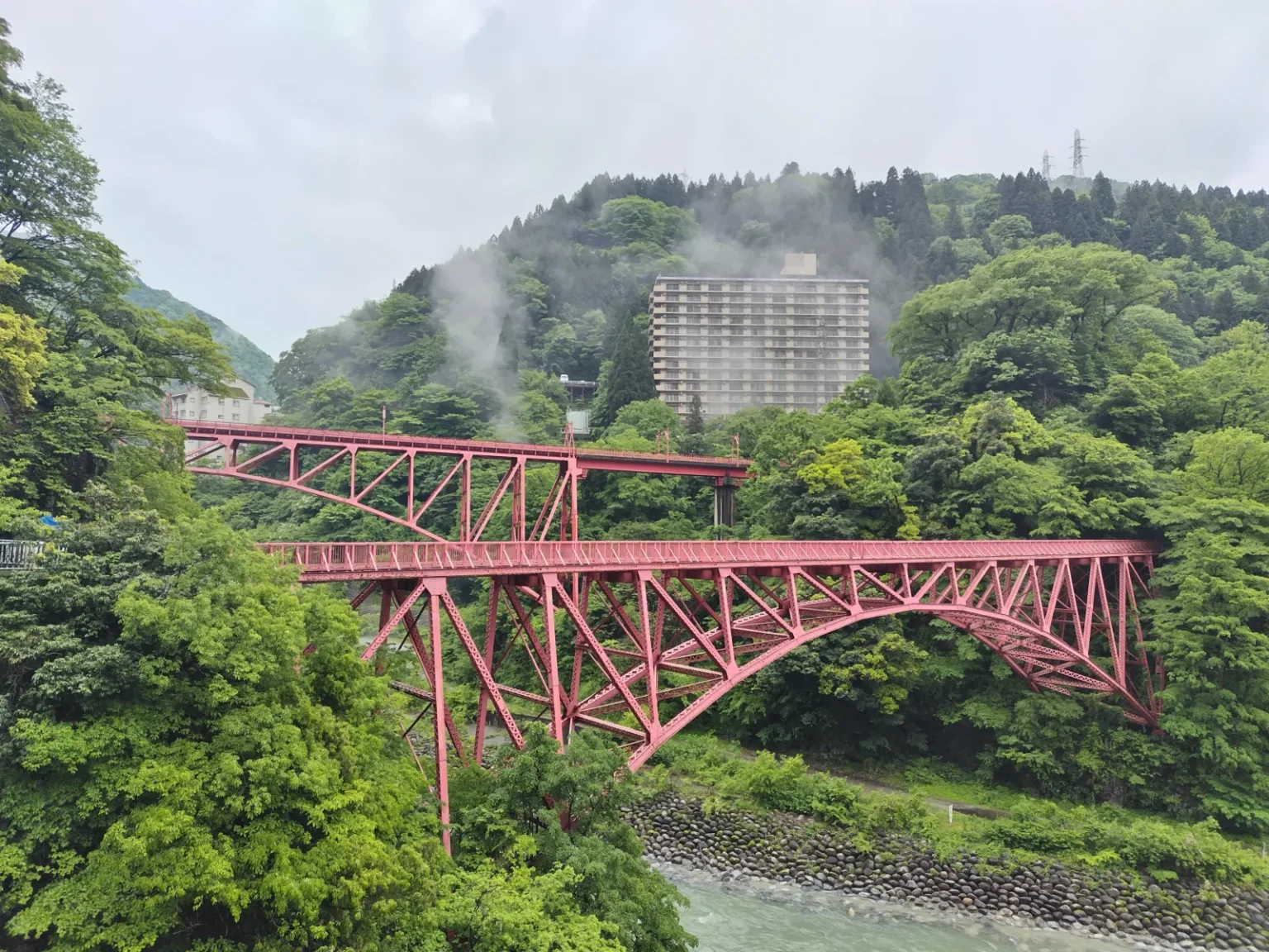 Two red steel bridges, one above the other, crossing a river between green wooded slopes, with a tall hotel building and drifting steam on the hillside behind