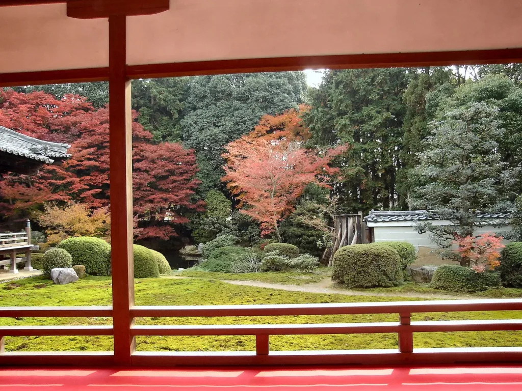 View from a red-floored temple veranda through red wooden pillars and a low rail onto a flat moss lawn with clipped round shrubs, a dark wall of evergreens, and maples turning red and orange.