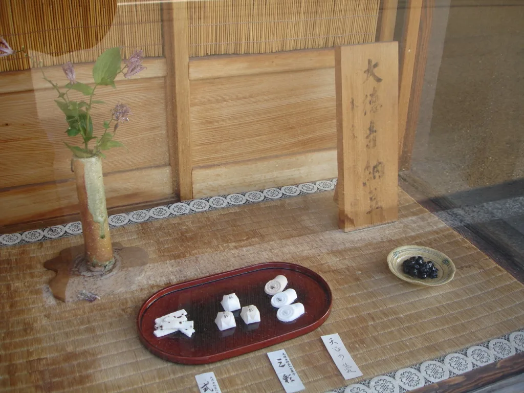 A shop window with a tatami floor: a tall wooden board brush-written with the characters 大徳寺納豆, a small ceramic dish of black, wrinkled beans to its right, a red lacquer tray of small white sweets with paper name cards in front, and a stem of flowers in a pottery vase at the left.