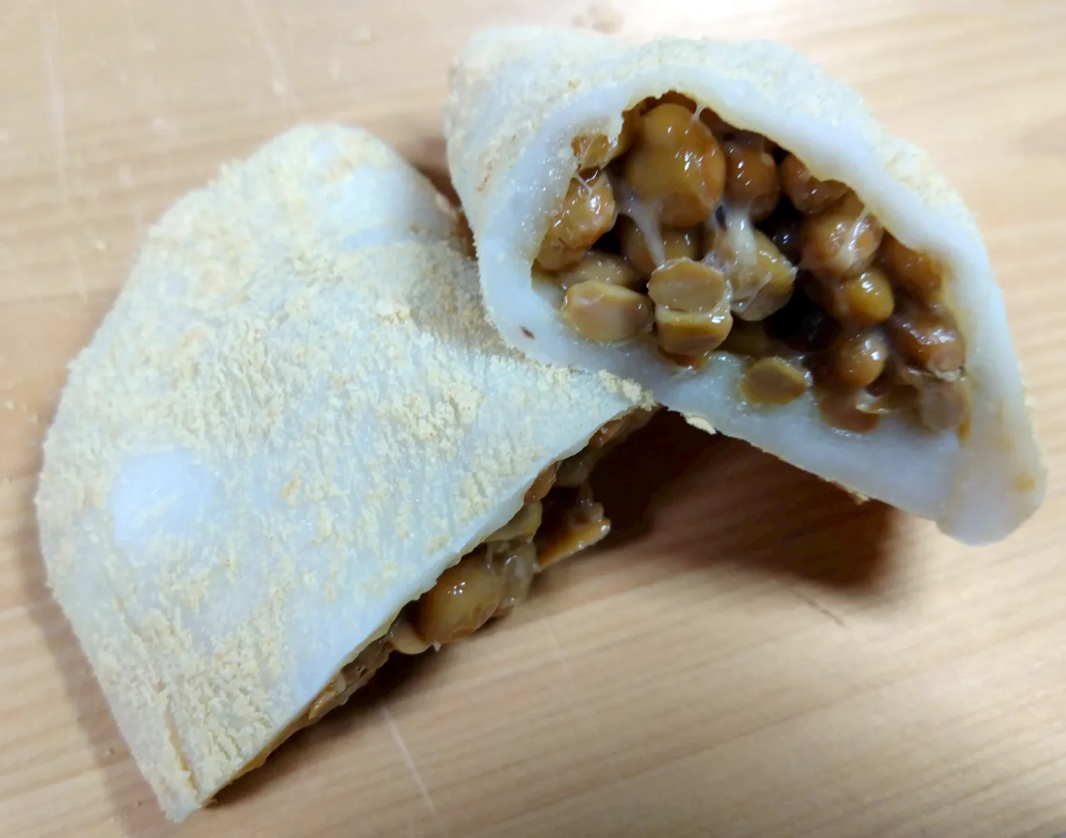 One soft white rice cake dusted with pale yellow kinako and cut in two on a wooden surface, the right-hand piece turned to show a filling of whole brown natto beans with a few sticky threads.