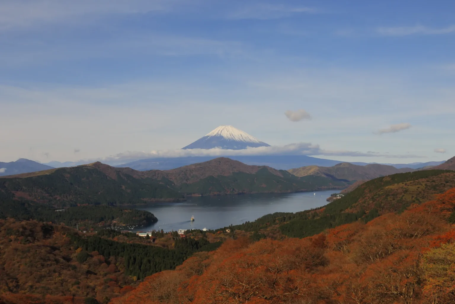 Snow-capped Mount Fuji above a band of cloud, with Lake Ashi below between forested ridges and red-brown autumn slopes in the foreground
