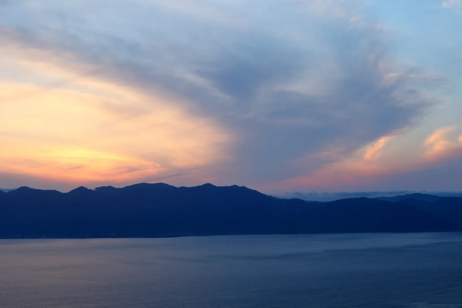 An orange and blue evening sky over the dark ridge of the Hira mountains and Lake Biwa, seen from the east shore