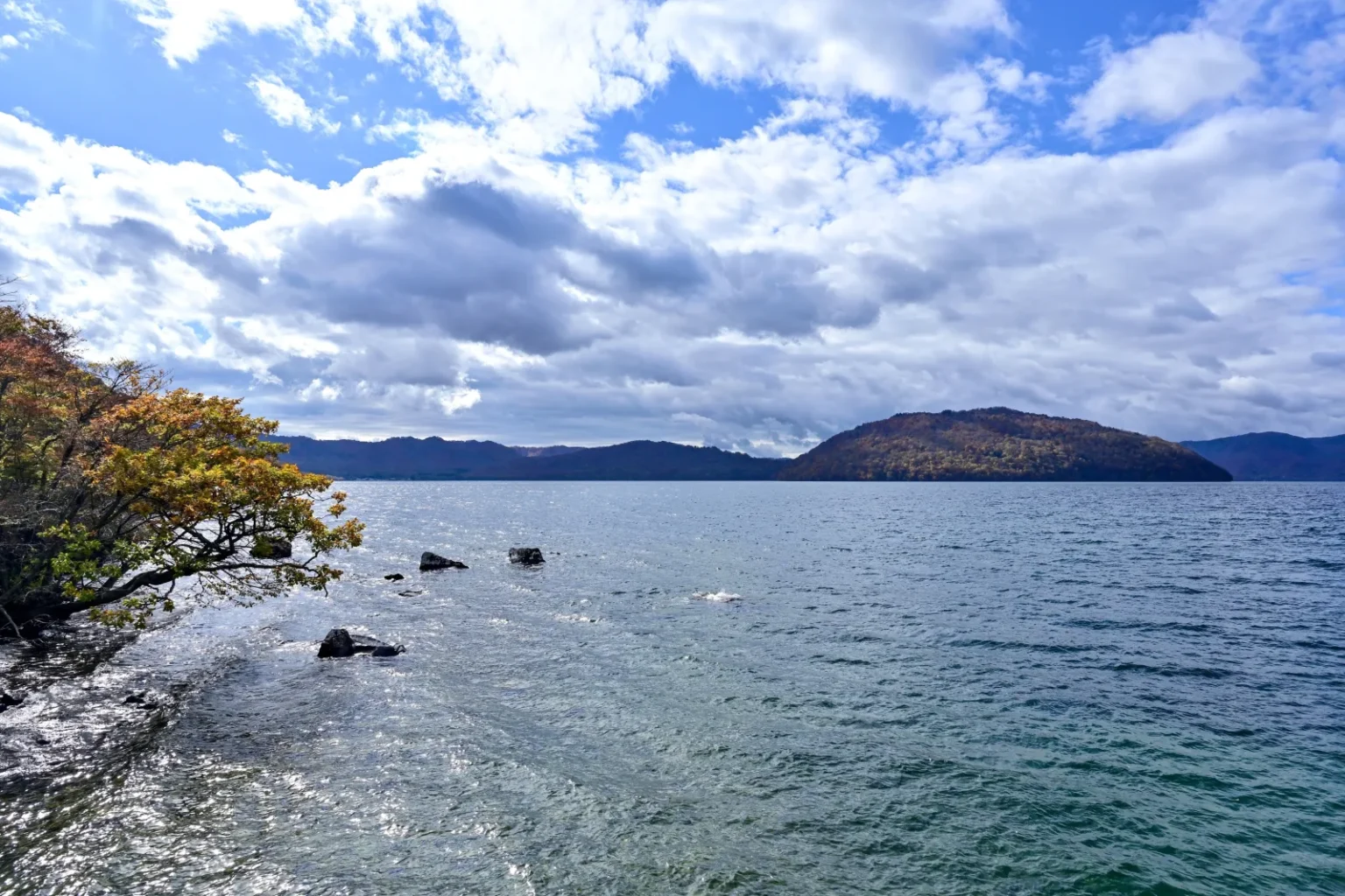 Lake Towada seen from the shore at Nenokuchi, with an autumn tree at the left edge, rocks in the water and a wooded headland under cloud