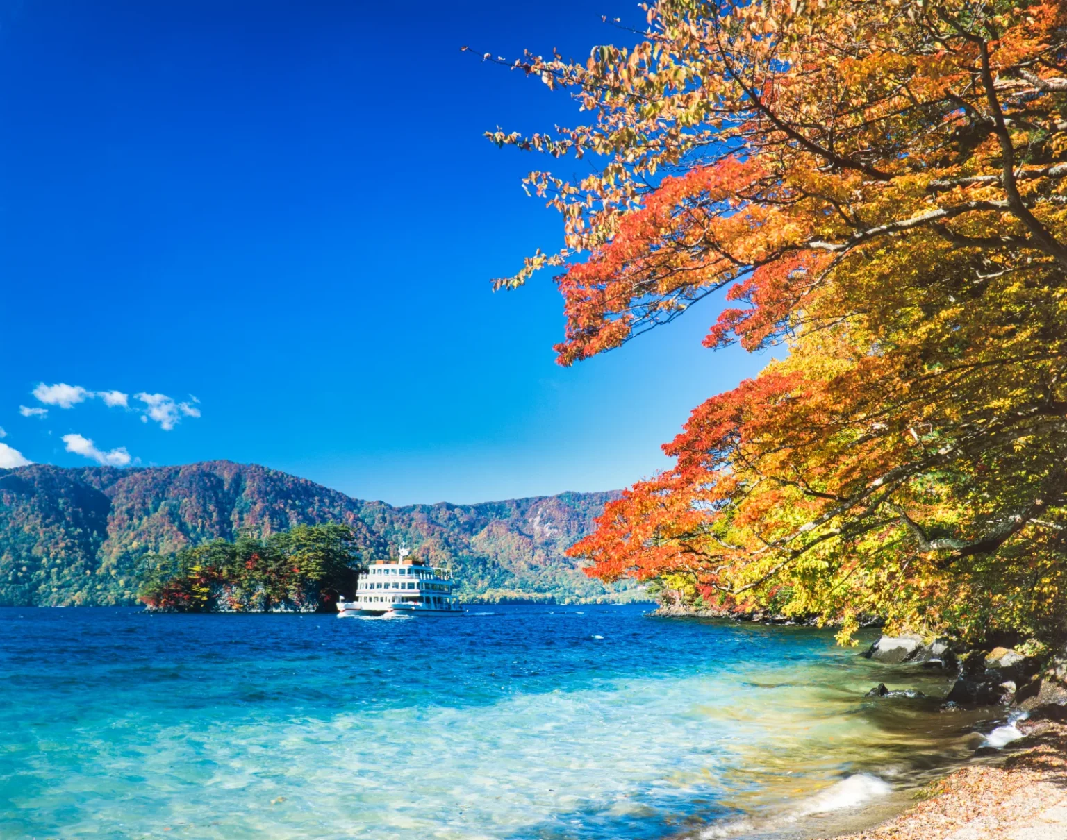 A white sightseeing boat on clear blue Lake Towada, with red and orange autumn leaves overhanging the shore and wooded hills across the water