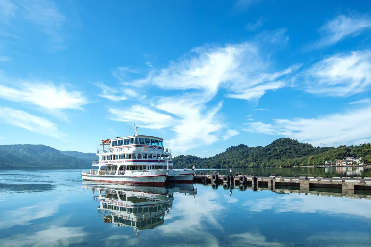 A white multi-deck sightseeing boat moored at the Yasumiya pier on Lake Towada, reflected in calm water under a blue sky with green hills behind