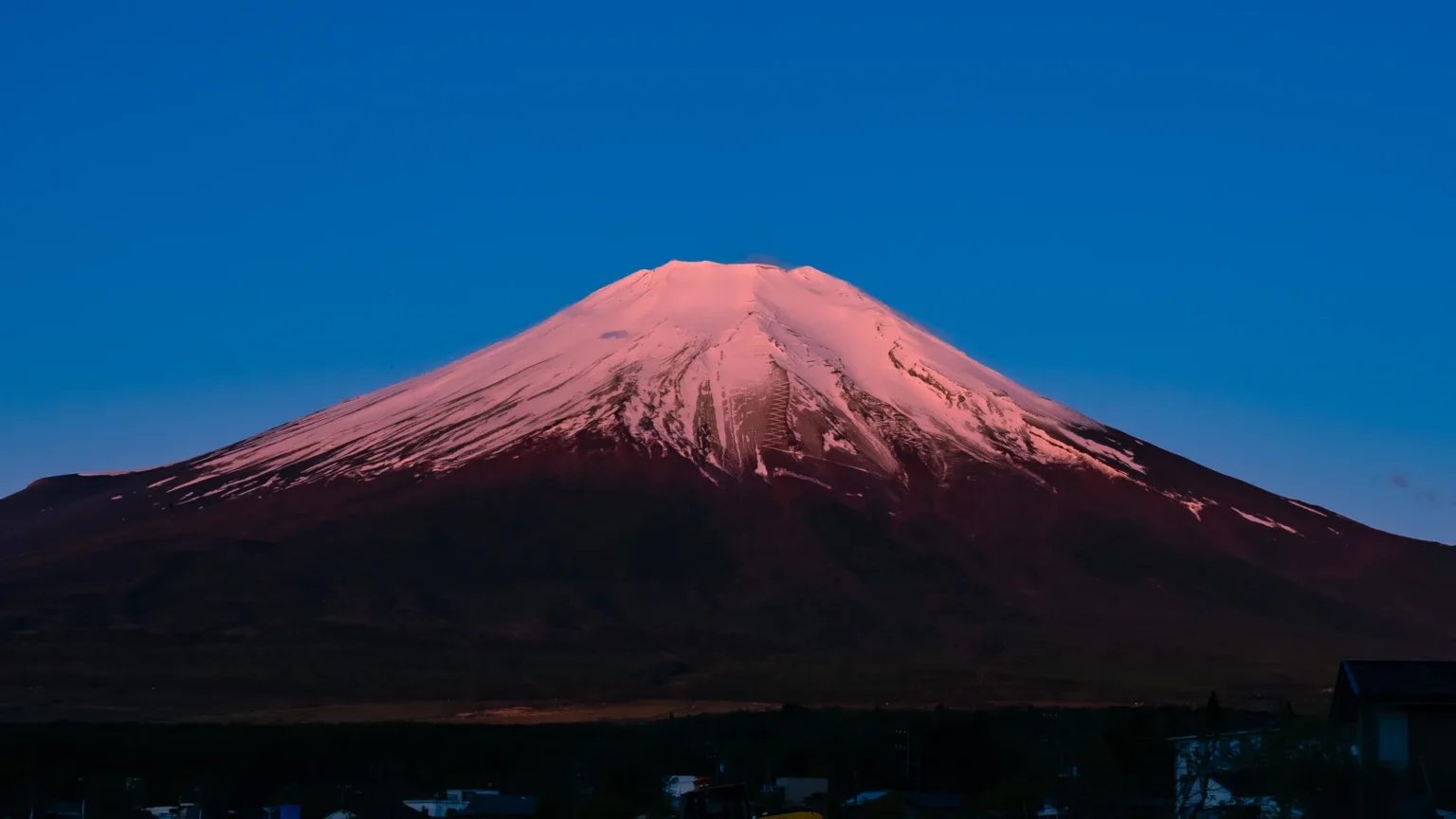The snow-covered summit of Mt Fuji lit pink by the first light of the morning under a deep blue sky, with the lower slopes still dark, photographed from Lake Yamanaka at 4:48 a.m. on 2 May 2026