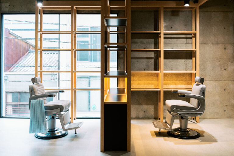 Two barber chairs facing tall windows framed in pale wood, with concrete walls