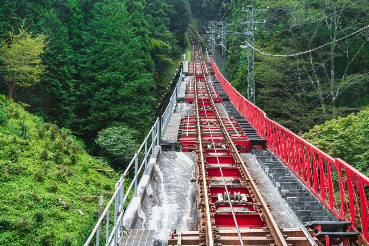 The steep track of the Mount Mitake cable car climbing through dense green forest on a red steel bridge in Ome, Tokyo, with the haul cable running between the rails and a long service stairway beside it