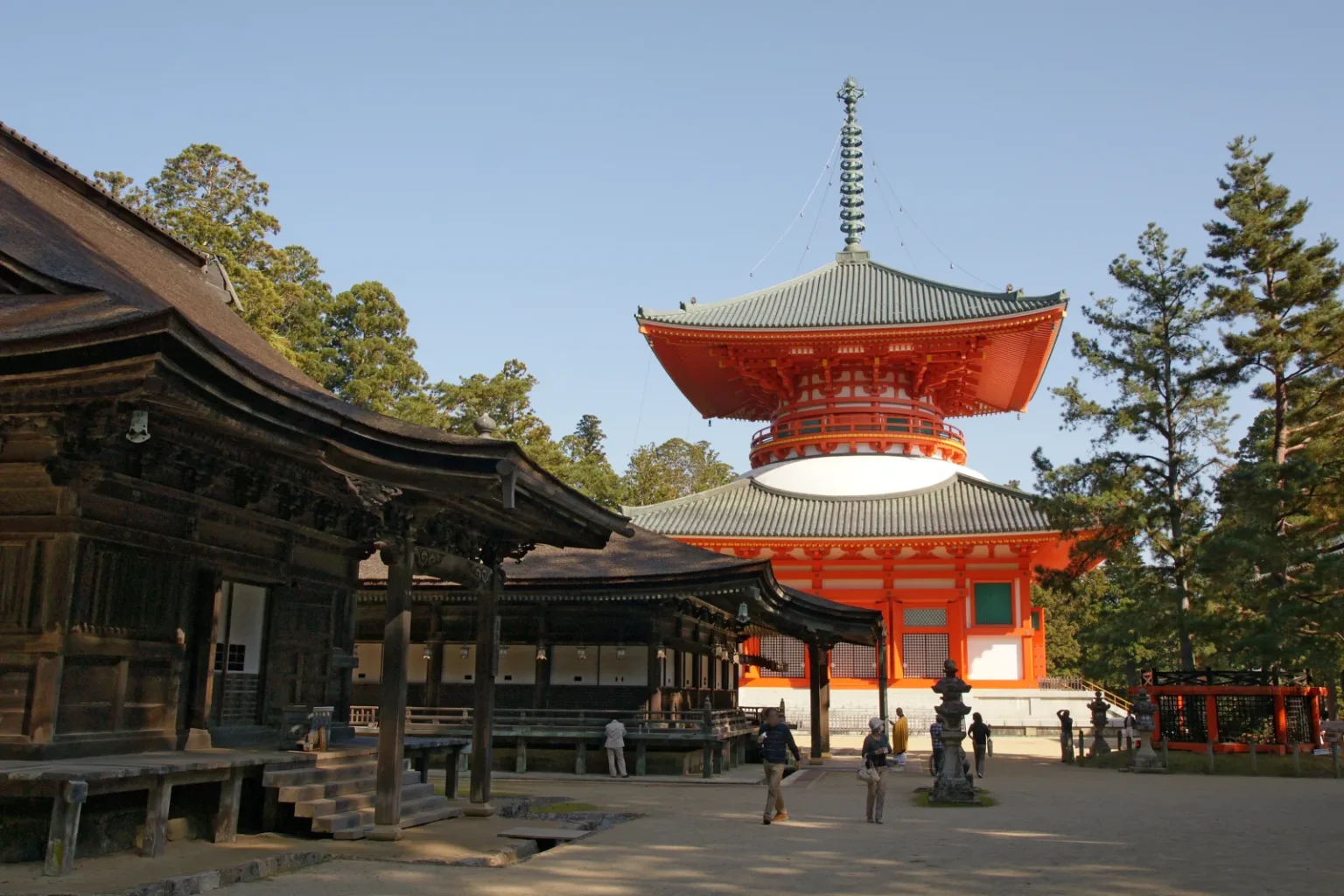 The orange-red Konpon Daito pagoda at Danjo Garan with a dark temple hall on the left and visitors walking across the courtyard