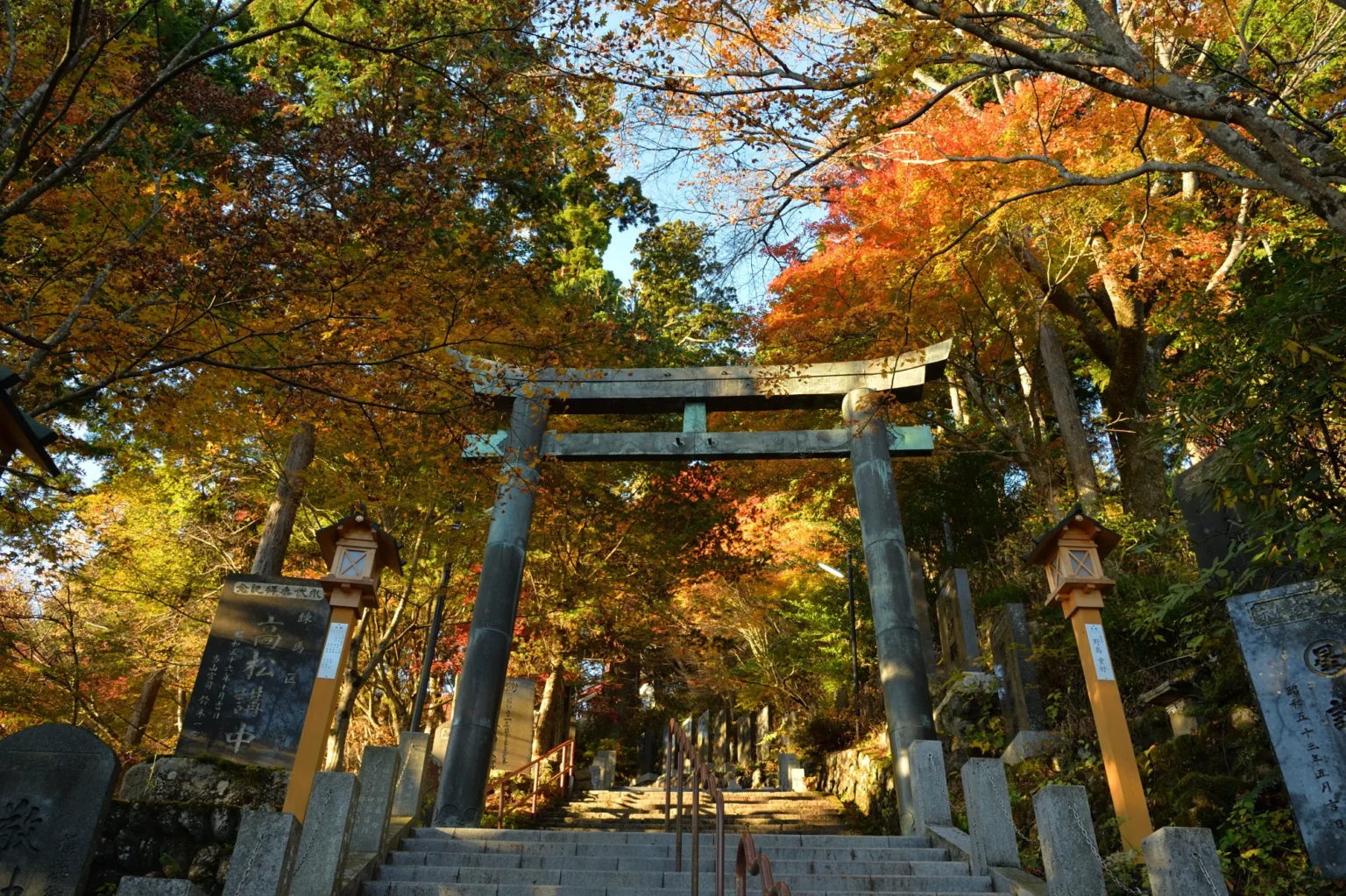 A green-patinated metal torii gate over the stone stairway to Musashi-Mitake Shrine on Mount Mitake, Tokyo, framed by orange and red autumn maples, with wooden lanterns and carved stone markers on both sides