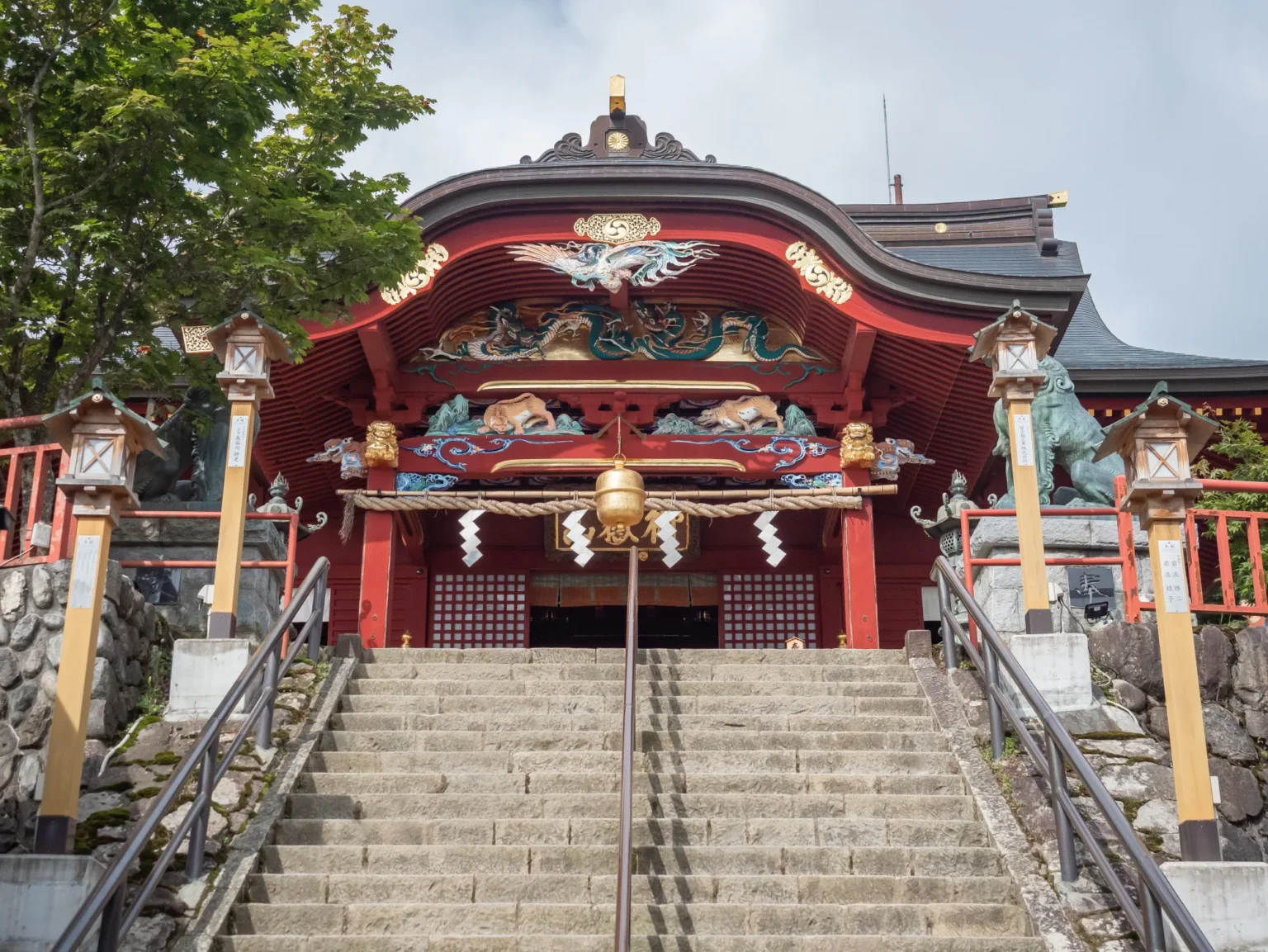 The red-lacquered worship hall of Musashi Mitake Shrine on Mount Mitake, Tokyo, with carved dragon and bird panels under the curved eaves and a large bell hung from a thick straw rope with white paper streamers, at the top of a long stone stairway