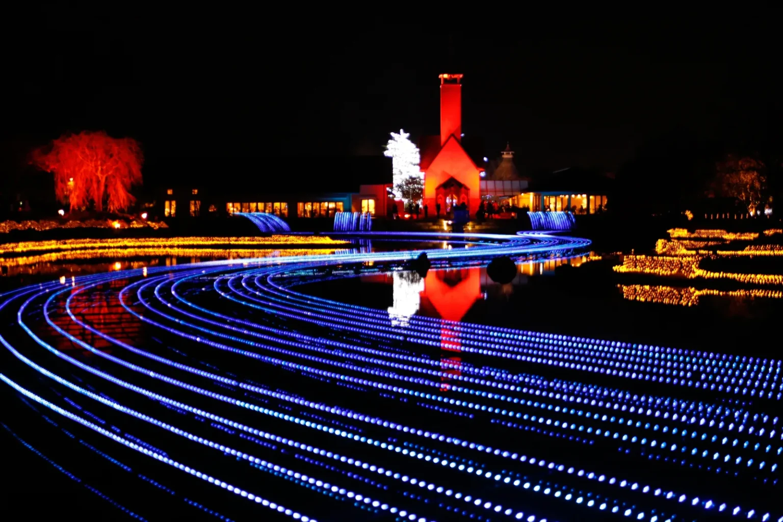 Curving rows of blue lights across the water at Nabana no Sato at night, with a lit red-brick building and a white-lit tree behind