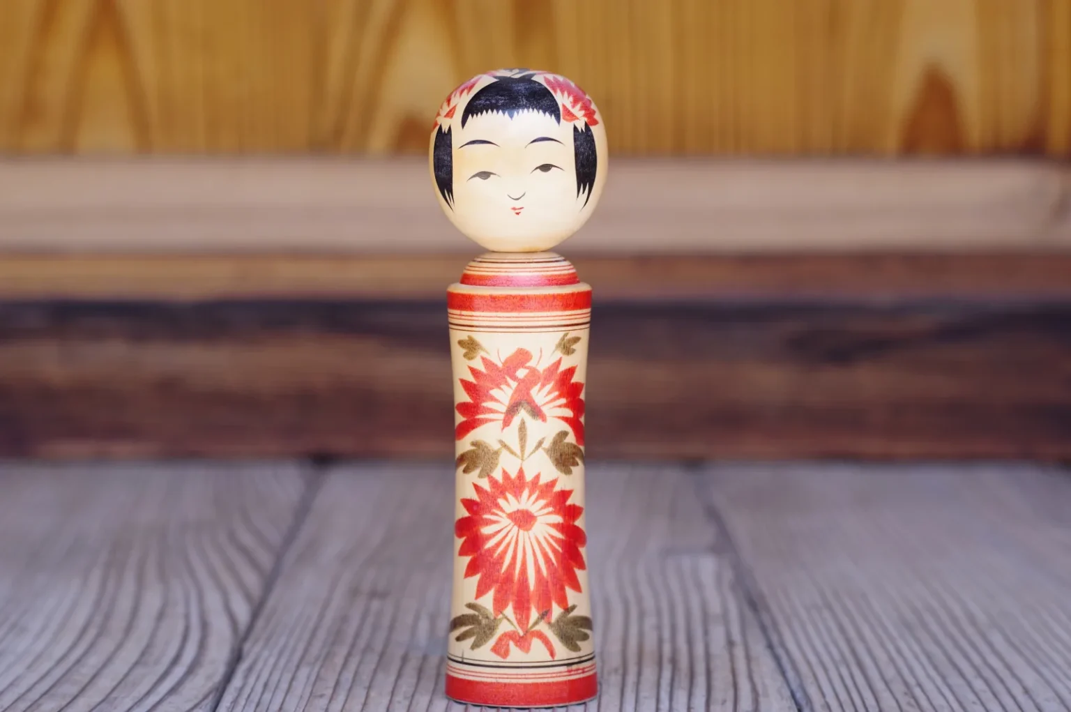 A single Naruko kokeshi doll with a painted face and red chrysanthemum pattern, standing on a wooden floor