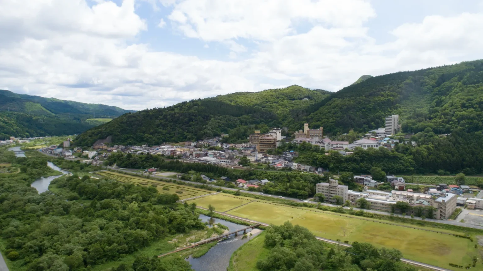 Aerial view of Naruko Onsen town: hotels and houses at the foot of wooded hills, with a river and fields in front
