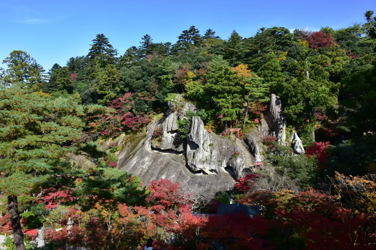 Pale weathered rock formations with small shrines, surrounded by pines and red autumn trees