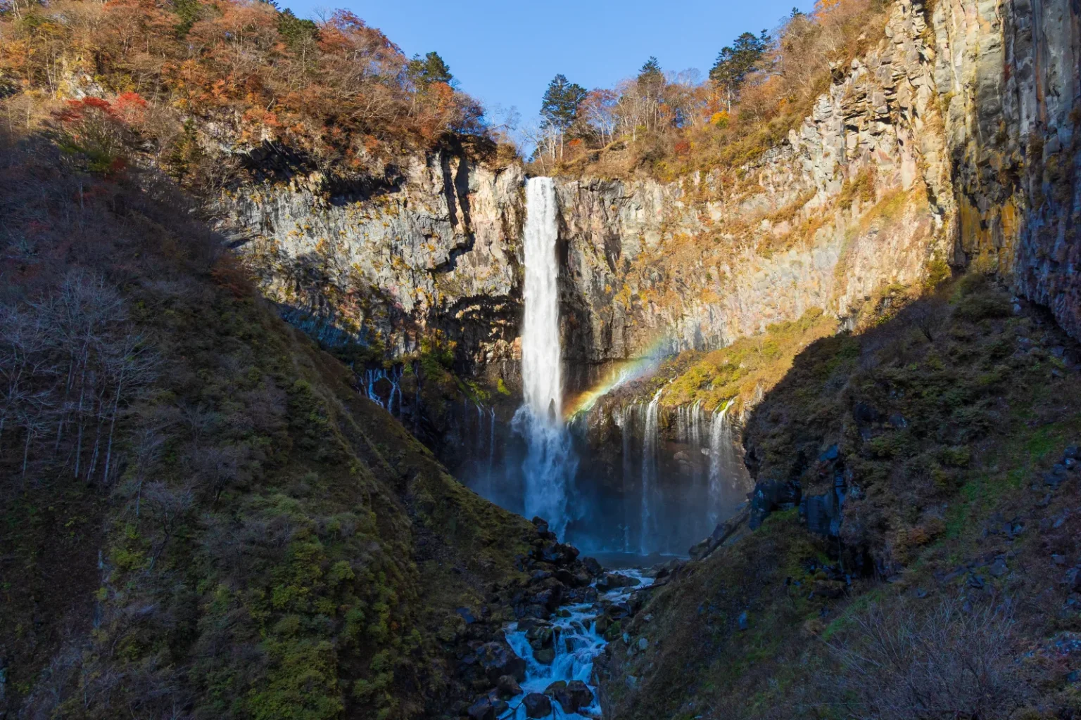 Kegon Falls in autumn, a tall single stream of water dropping down a sheer rock cliff with a faint rainbow at its base
