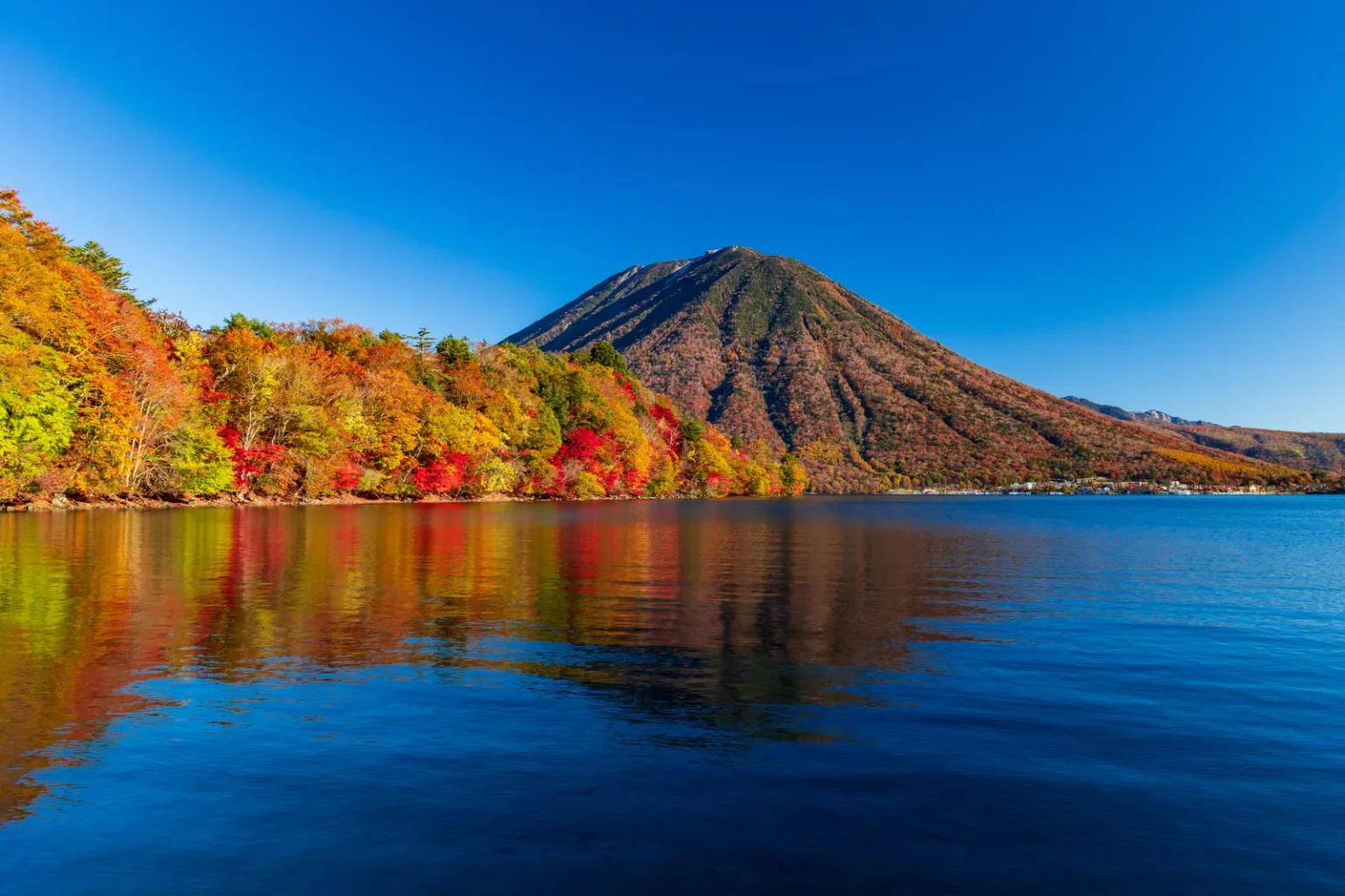 Lake Chuzenji in autumn with red and orange trees along the shore and the cone of Mt Nantai behind
