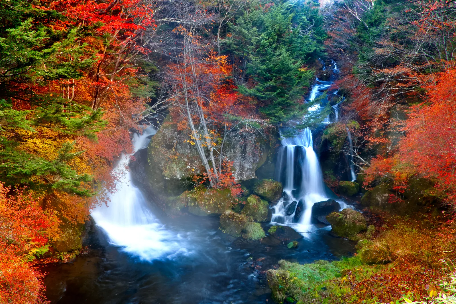 Ryuzu Falls in Oku-Nikko in autumn, white water splitting around a mossy rock between red, orange and yellow trees