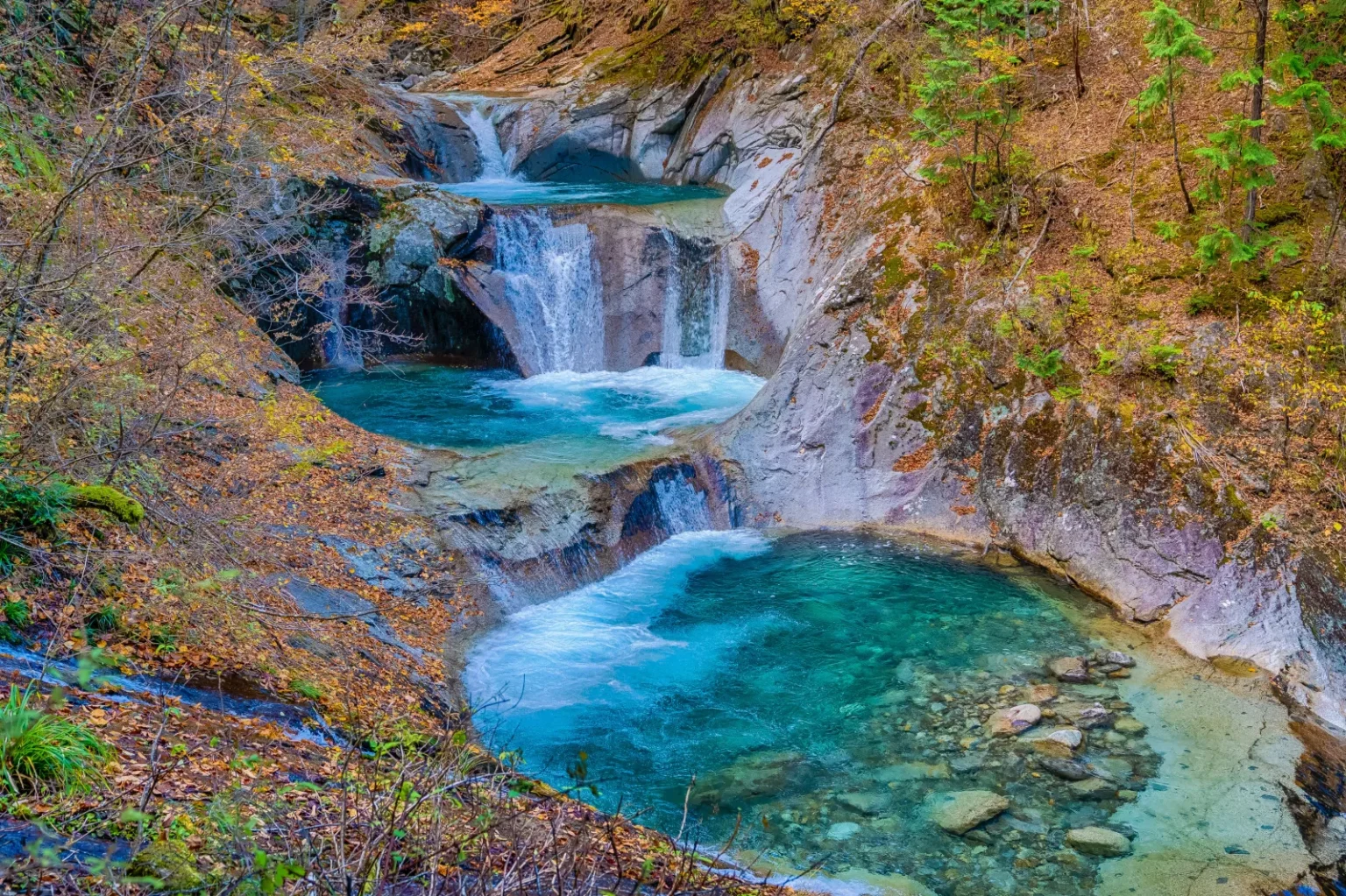 A series of small waterfalls dropping into clear blue-green pools between pale rock walls, with autumn leaves around