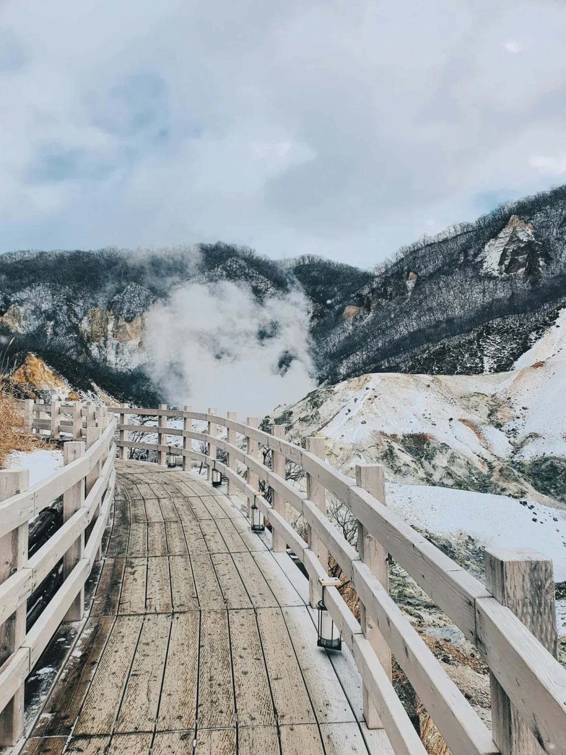 A curving wooden boardwalk with pale railings and small lanterns crossing snow-covered ground in Jigokudani, Noboribetsu, with steam rising against dark wooded slopes and a cloudy sky