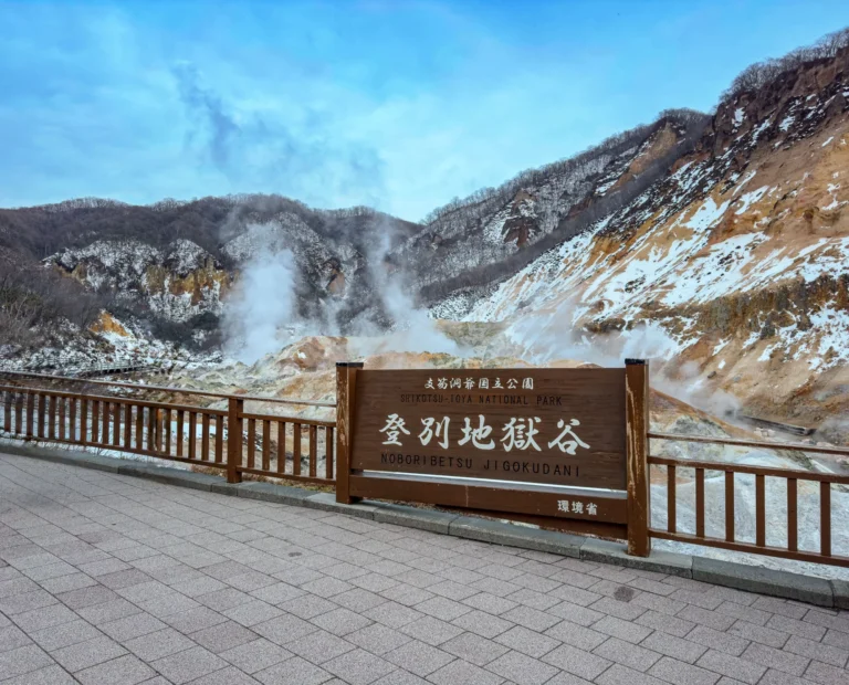 A brown wooden sign reading Shikotsu-Toya National Park, Noboribetsu Jigokudani, Ministry of the Environment, on a paved promenade with a wooden railing, in front of a steaming crater valley with orange and white rock and a light covering of snow on the slopes